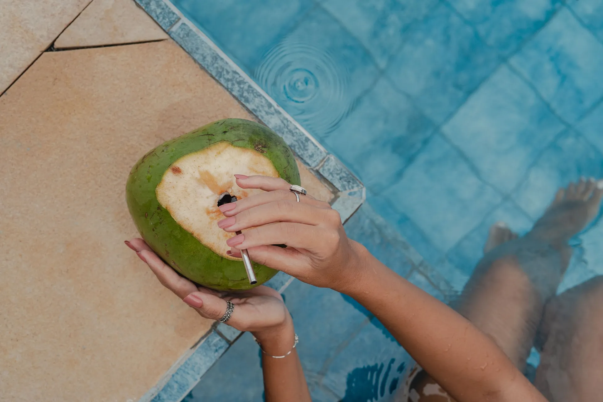 Mãos seguram um coco verde na borda da piscina do Chocolate Hotel, com os pés dentro da água