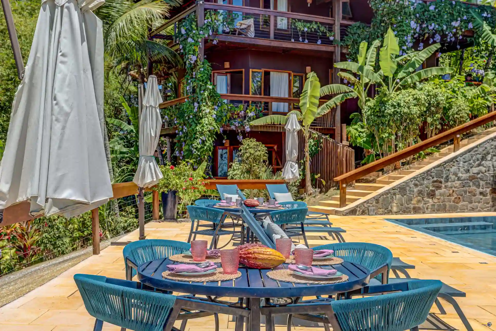 As mesas do deck da piscina do Chocolate Hotel, com os guarda-sóis e o prédio de madeira entre as bananeiras