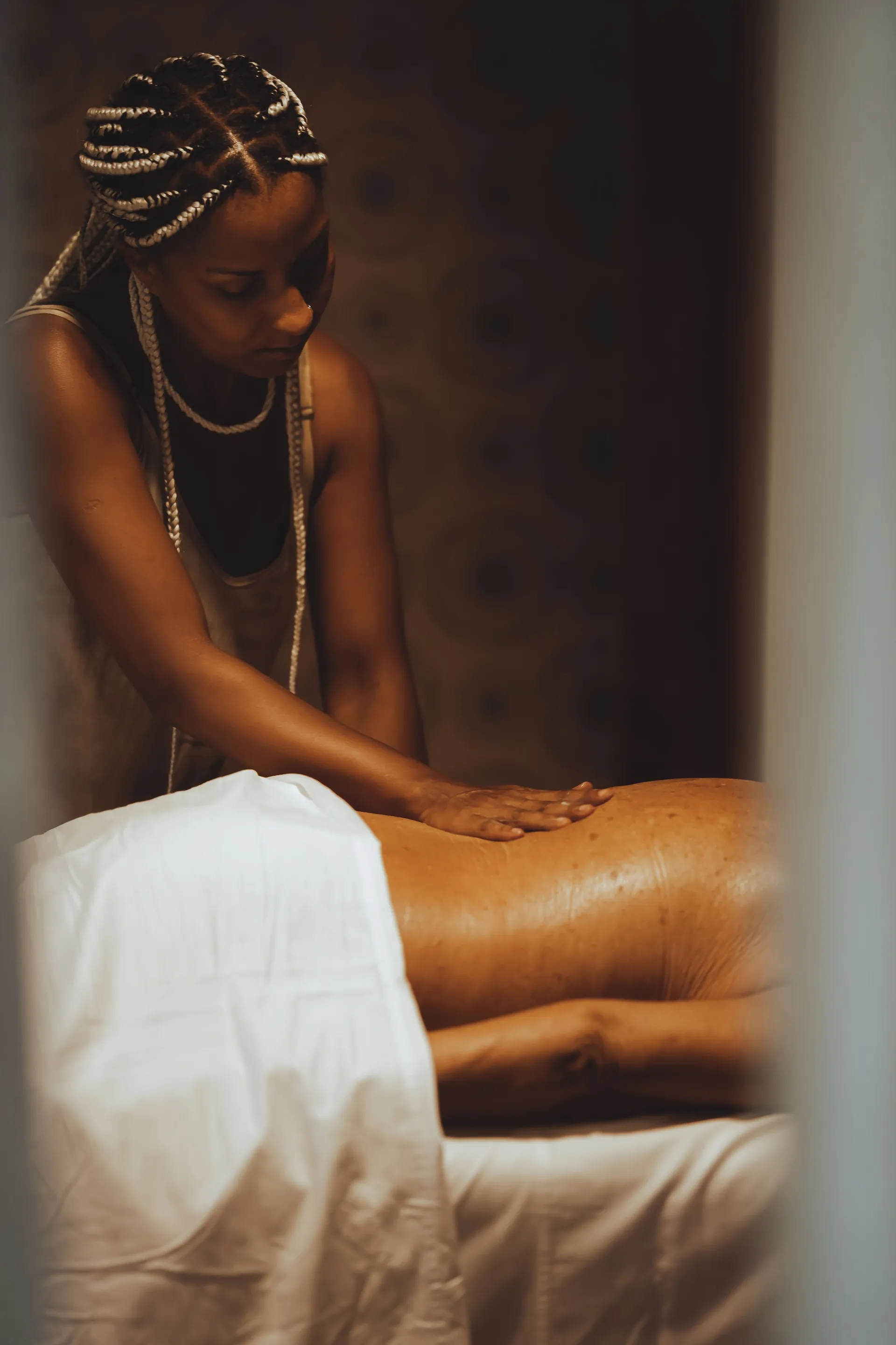 Massagem em curso: as mãos da profissional nas costas de quem recebe, lençol branco e parede de madeira escura