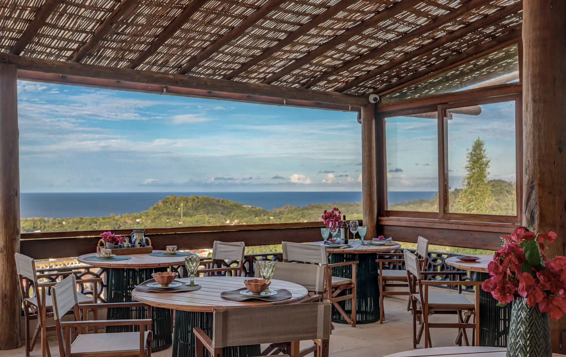 Salão do restaurante Nibs sob o forro de bambu, mesas postas de madeira e a paisagem de Itacaré aberta ao fundo: a mata, o mar e o horizonte