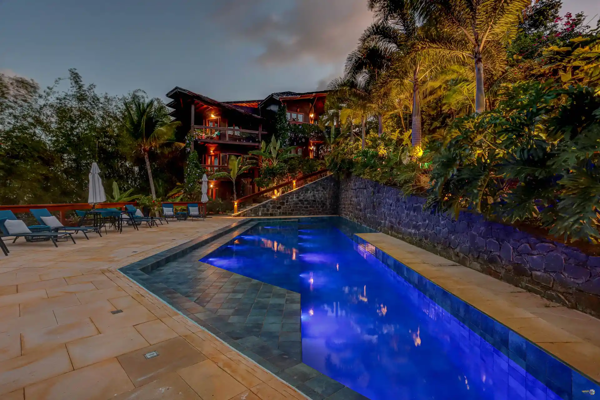 A piscina do Chocolate Hotel ao anoitecer, com o hotel iluminado por dentro e espreguiçadeiras vazias
