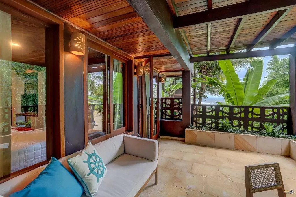The covered balcony of a Tulha suite, with a sofa and the sea showing through the trees